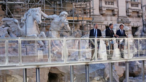 Drei Männer in Anzügen stehen vor dem Trevi-Brunnen in Rom, der sich im Umbau befindet.