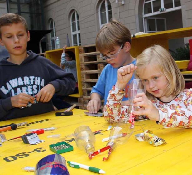 Drei Kinder basteln an einem gelben Tisch mit Werkzeugen und Recyclingmaterialien.