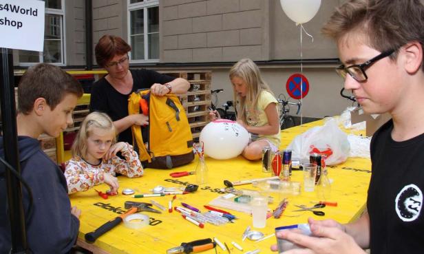 Kinder und eine Frau sitzen an einem gelben Tisch und basteln mit verschiedenen Materialien.
