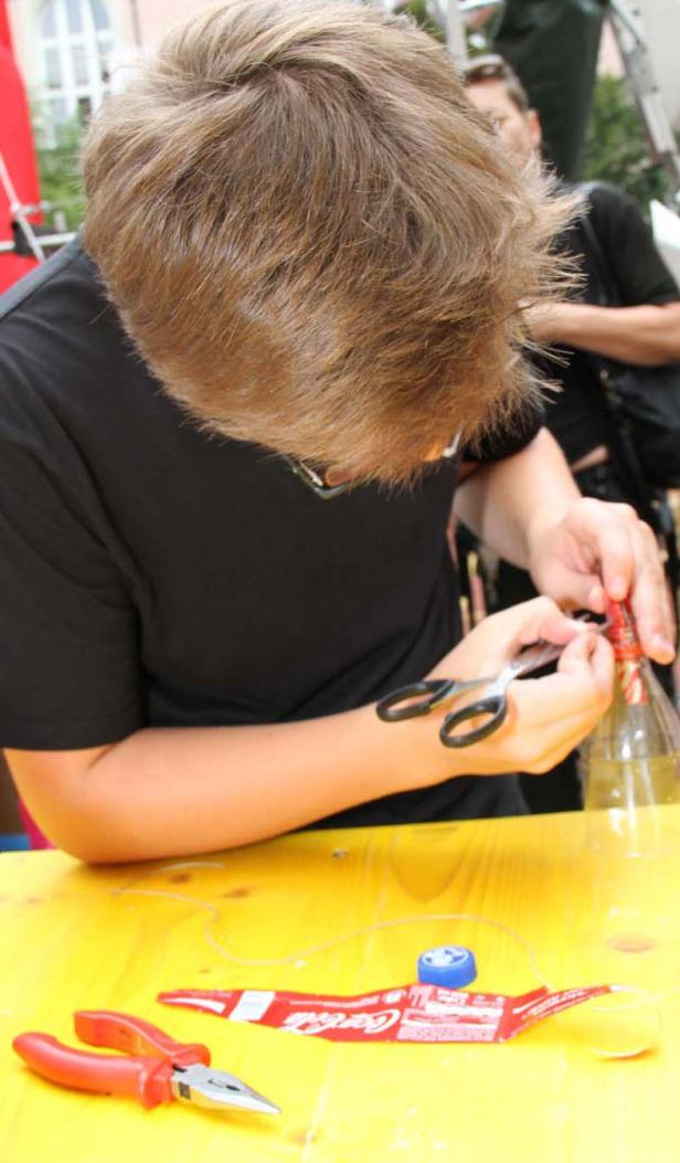Eine Person bastelt mit einer Plastikflasche, einer Schere und einer Zange an einem gelben Tisch.