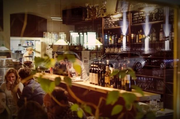 Blick durch ein Fenster in eine Bar mit Gästen und einem Barkeeper hinter der Theke.