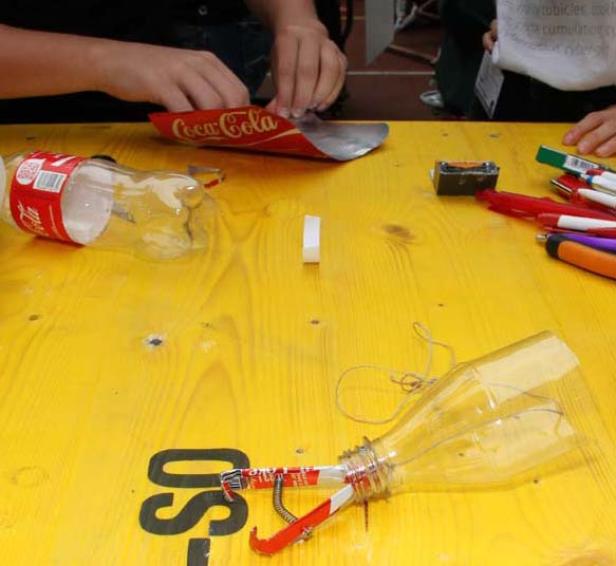 Kinder basteln mit Plastikflaschen und Coca-Cola-Verpackungen auf einem gelben Tisch.