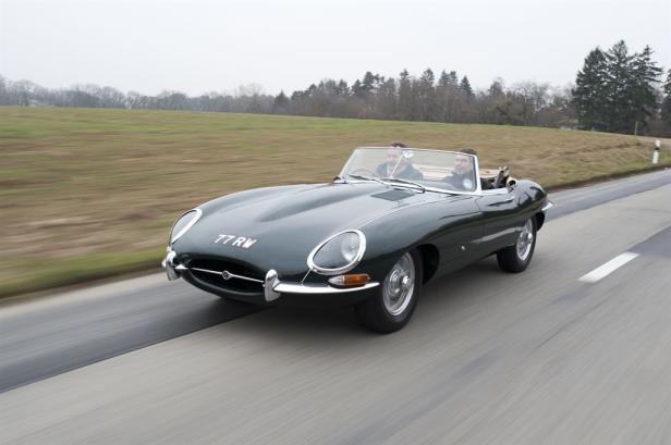 Ein grüner Jaguar E-Type Roadster fährt auf einer Landstraße.
