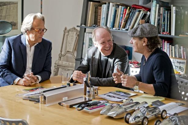 Drei Männer sitzen an einem Tisch mit Modellautos und diskutieren.
