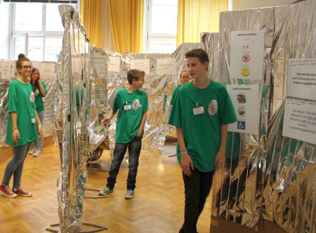 Schüler in grünen T-Shirts stehen in einem Raum mit Installationen zum Thema Armut.