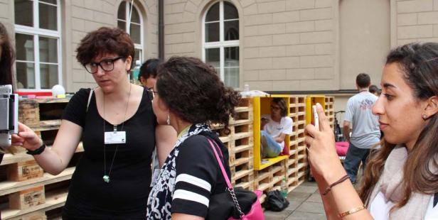 Zwei Frauen interagieren mit einer Installation aus Holzpaletten, während eine dritte ein Foto macht. Eine Person sitzt in einem gelben Rahmen aus Paletten.