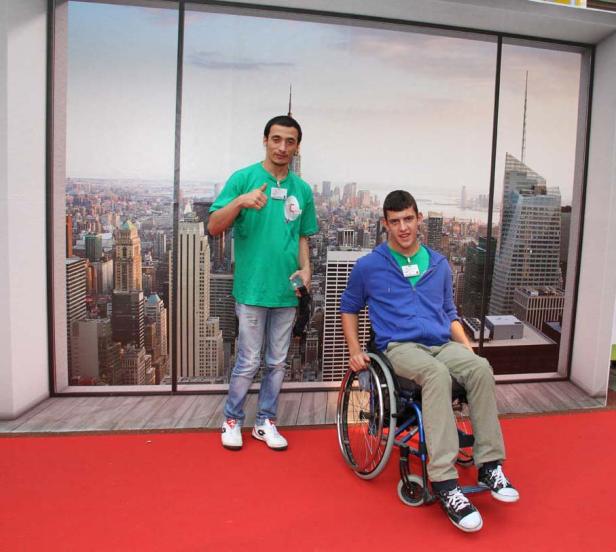 Zwei junge Männer posieren vor einem New Yorker Skyline-Hintergrund. Einer sitzt im Rollstuhl.