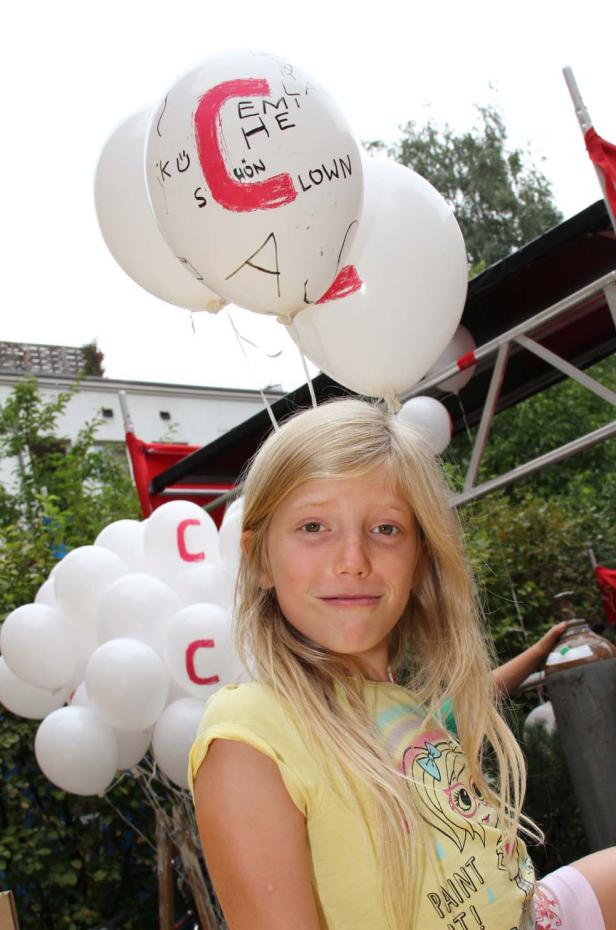 Ein blondes Mädchen steht vor einem Wagen mit weißen Luftballons, die mit roter Farbe bemalt sind.