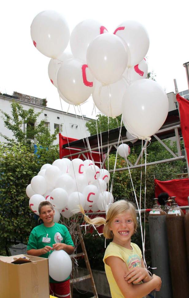 Zwei Kinder stehen vor einem Stand mit weißen Luftballons mit rotem „C“.
