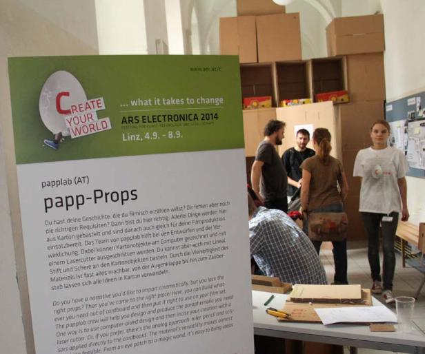 Ein Stand von Ars Electronica 2014 mit dem Thema Papp-Props und drei Personen im Hintergrund.