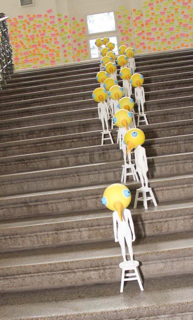 Eine Treppe hinauf stehen Figuren mit gelben Vogelkopf-Masken auf kleinen Hockern.