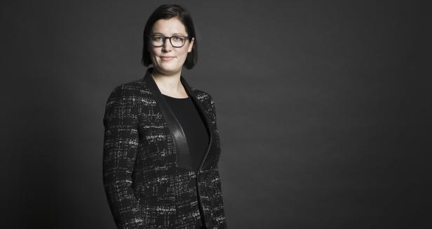 Eine Frau mit Brille und Blazer vor einem grauen Hintergrund.