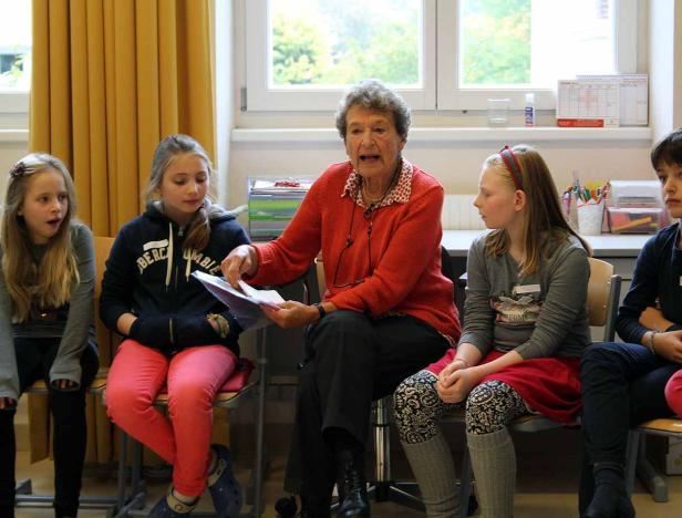 Eine ältere Frau liest einer Gruppe von Kindern in einem Klassenzimmer vor.