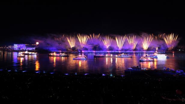 Mehrere beleuchtete Boote fahren auf einem Fluss unter einem Feuerwerk.