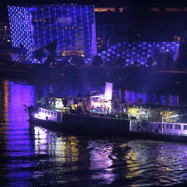Ein beleuchtetes Schiff fährt auf einem Fluss vor einem blau beleuchteten Gebäude.