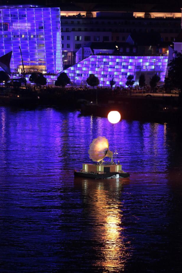 Eine schwimmende Plattform mit einem Grammophon und einem leuchtenden Ballon auf einem Fluss.
