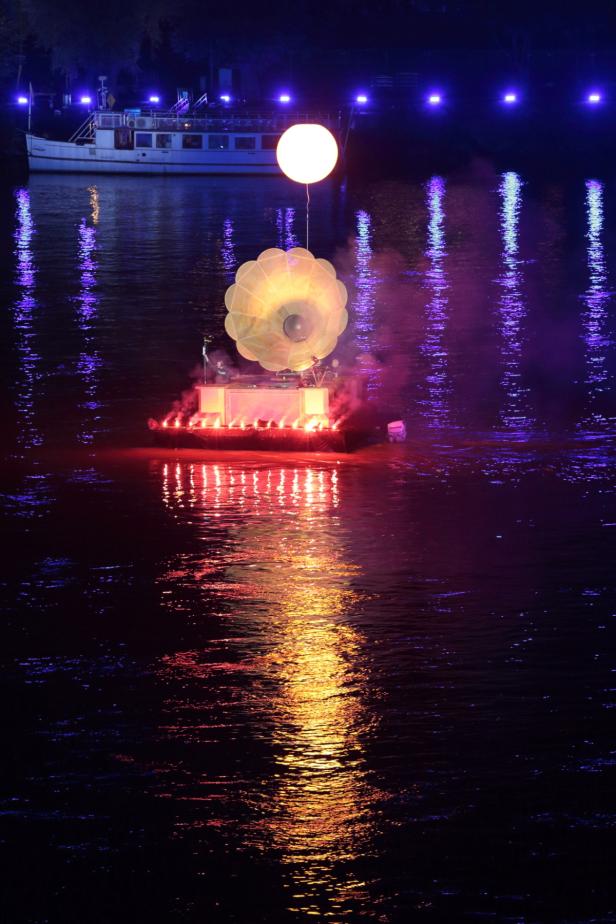 Eine beleuchtete Bühne schwimmt auf einem Fluss, mit einem Ballon und einem Schiff im Hintergrund.