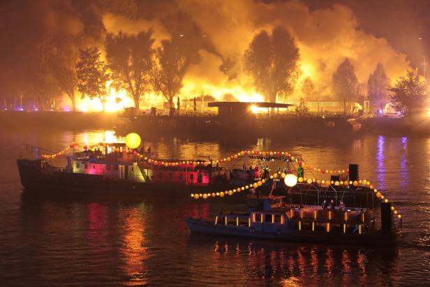 Beleuchtete Schiffe fahren auf einem Fluss, im Hintergrund ein Feuerwerk.