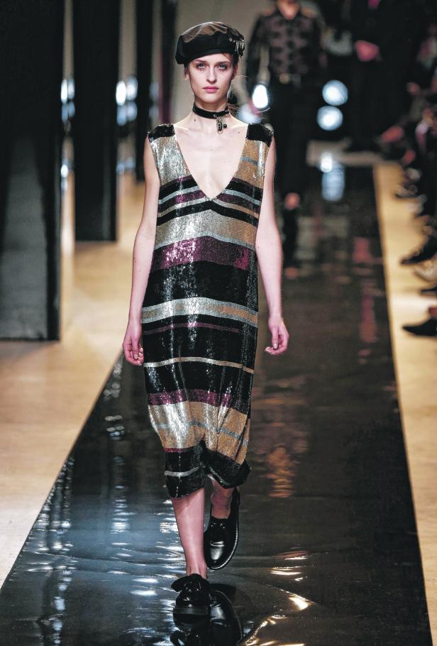 Eine Frau mit Barett und Paillettenkleid läuft über einen glänzenden Laufsteg.