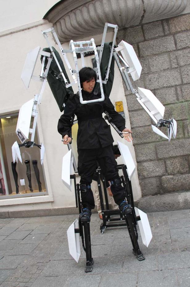 Eine Person in einem schwarzen Anzug trägt ein großes, weißes Exoskelett auf der Straße.