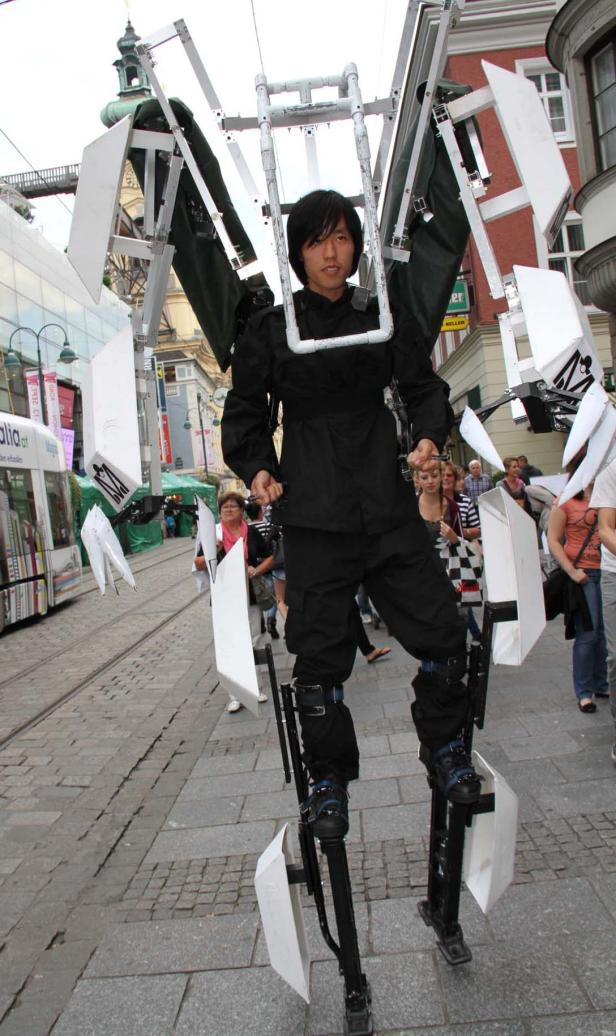 Ein Mann in einem schwarzen Anzug und Stelzen mit weißen Paneelen steht auf einer belebten Straße.