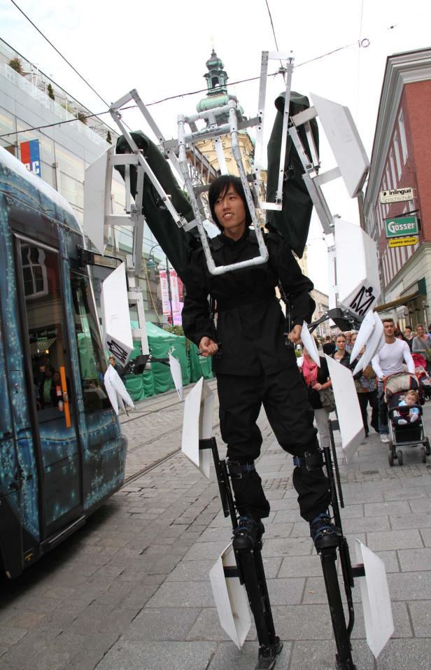 Ein Mann in einer Performance-Montur steht auf Stelzen in einer Stadt.