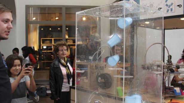 Zuschauer beobachten eine Maschine mit fallenden blauen Bechern in einem transparenten Gehäuse.