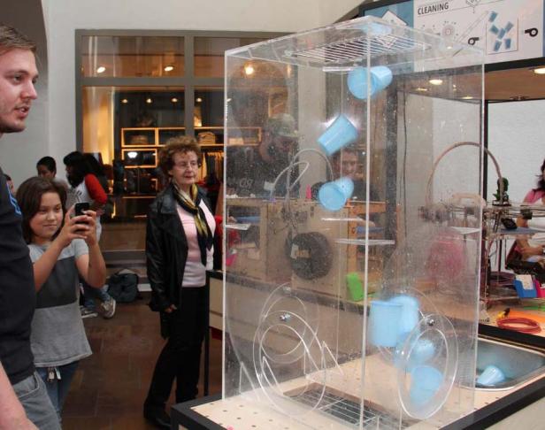 Zuschauer beobachten eine Maschine mit blauen Bechern in einem transparenten Gehäuse.