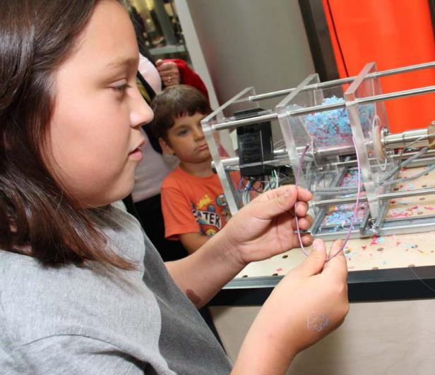 Ein Mädchen betrachtet eine Maschine mit bunten Konfetti-Schnipseln, während ein Junge im Hintergrund zusieht.