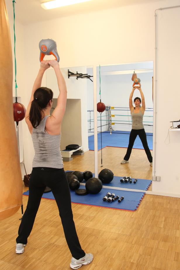 Eine Frau trainiert in einem Fitnessstudio mit einer 8-kg-Kettlebell über dem Kopf.