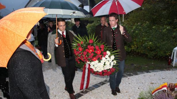 Zwei Männer in Tracht tragen unter Regenschirmen einen großen Blumenkranz.