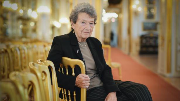 Eine ältere Frau sitzt in einem eleganten Saal auf einem goldenen Stuhl.
