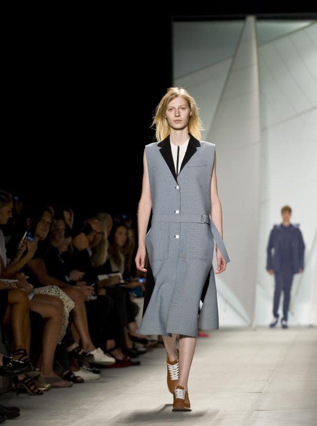 Eine blonde Frau läuft auf einem Laufsteg und präsentiert ein ärmelloses Kleid mit Blazer-Details.