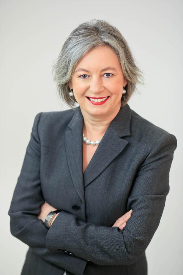 Eine Frau mit grauen Haaren, gekleidet in einem grauen Blazer, lächelt mit verschränkten Armen.