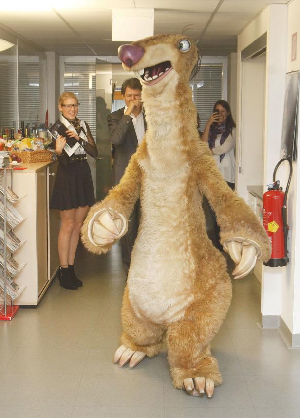 Ein Sid-Faultier-Kostüm steht in einem Büro, während Leute im Hintergrund zusehen.