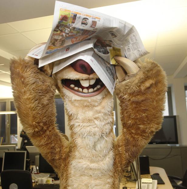 Ein Kostüm-Faultier hält Zeitungen über seinem Kopf.