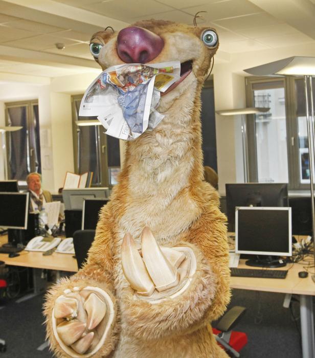 Ein Sid-Faultier-Kostüm hält Zeitungen im Mund und in den Händen in einem Büro.