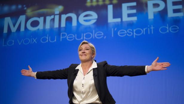 Marine Le Pen steht mit ausgestreckten Armen vor einem blauen Hintergrund.