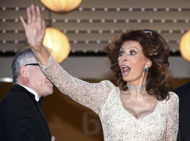 Sophia Loren winkt auf einer Veranstaltung mit einem Mann im Hintergrund.