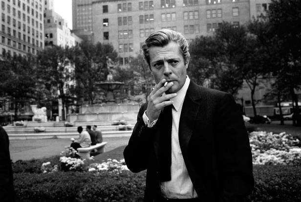 Schwarzweißaufnahme von William Holden, der in einem Park eine Zigarette raucht.