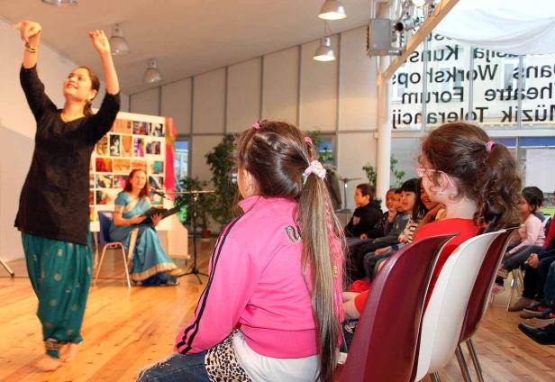 Eine Frau tanzt vor einem Publikum, während eine andere Frau Musik spielt.