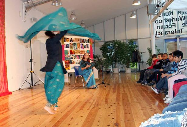 Eine Frau tanzt in traditioneller Kleidung vor einer Gruppe von Kindern, während eine andere Frau singt.