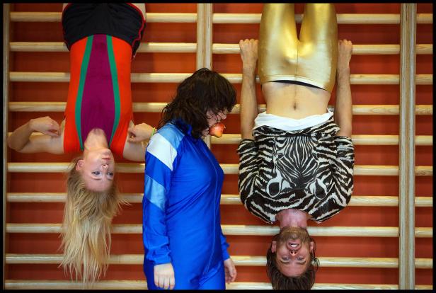 Zwei Personen hängen kopfüber an einer Sprossenwand, während eine Frau mit Apfel im Mund daneben steht.