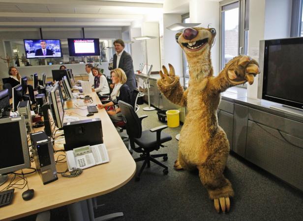 Ein Sid-Faultierkostüm steht in einem Büro mit mehreren Mitarbeitern.
