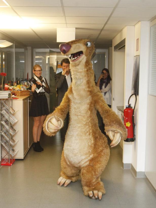 Ein Sid-Faultier-Kostüm steht in einem Büro, umgeben von Menschen.