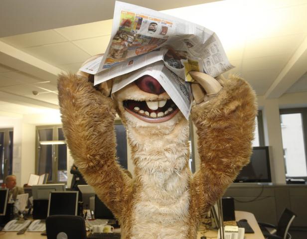 Ein Faultier-Kostüm hält sich Zeitungen über den Kopf.