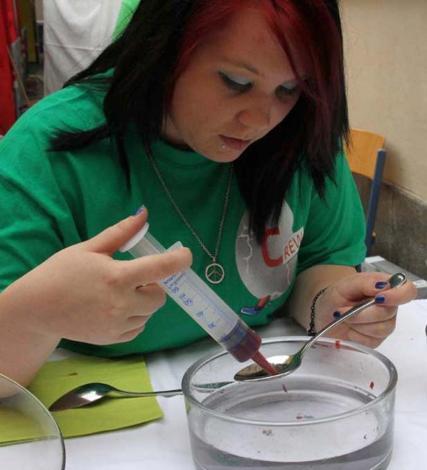 Eine junge Frau mit Piercing füllt mit einer Spritze rote Flüssigkeit auf einen Löffel.