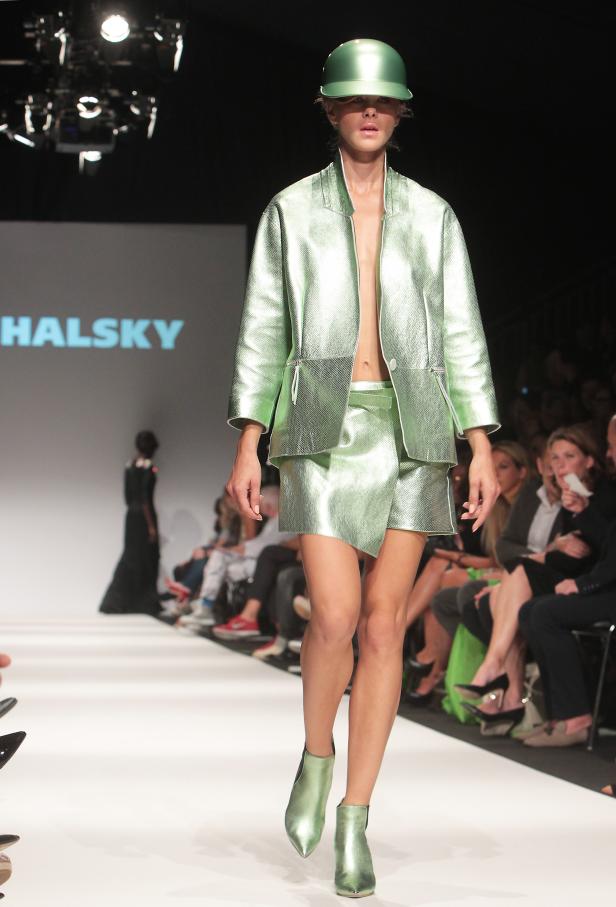 Eine Frau in einem glänzend grünen Ensemble mit passendem Hut auf einem Laufsteg.