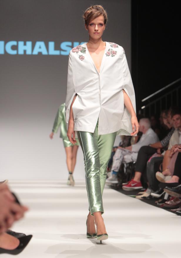 Eine Frau präsentiert ein weißes Oberteil mit Blumenapplikationen und glänzende, grüne Hosen auf dem Laufsteg.