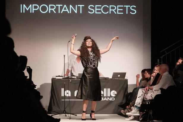Eine Sängerin mit lockigem Haar tritt vor dem Publikum mit dem Schriftzug „Important Secrets“ im Hintergrund auf.